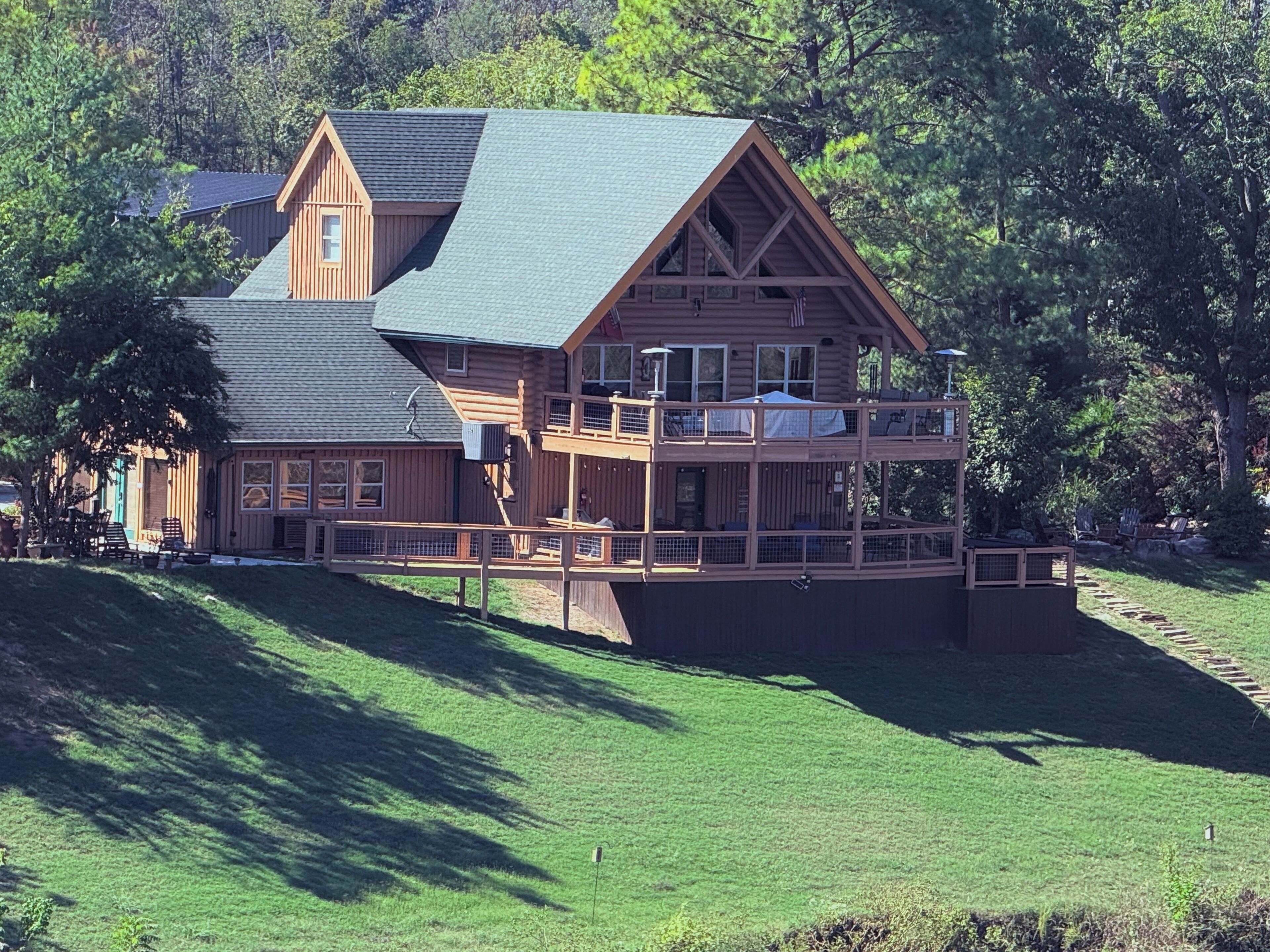 A large log cabin with a two-story deck, surrounded by lush greenery.