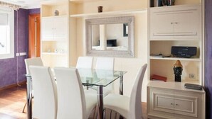 A dining area with a glass table and white chairs, a white cabinet with a mirror, and a bookshelf with books and a radio. - Spectacular Five Senses Mallorca Apartment (Barcelona)