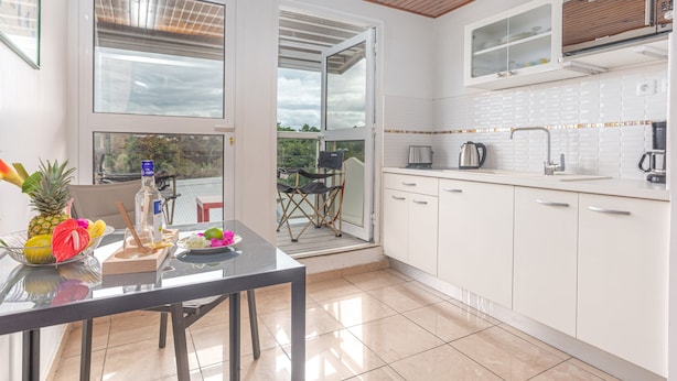 A modern kitchen with white cabinets, a dining table with a fruit bowl, and a view of the outdoors. - Studio 204 - Vanille - Proche plage (Les Trois-Îlets)