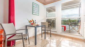 A room with a table, two chairs, a red curtain, a framed picture, and a view of the outdoors. - Studio 204 - Vanille - Proche plage (Les Trois-Îlets)