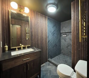 Un baño con tocador de madera, un espejo grande, ducha con patrón de mosaico de espiga y un inodoro.
