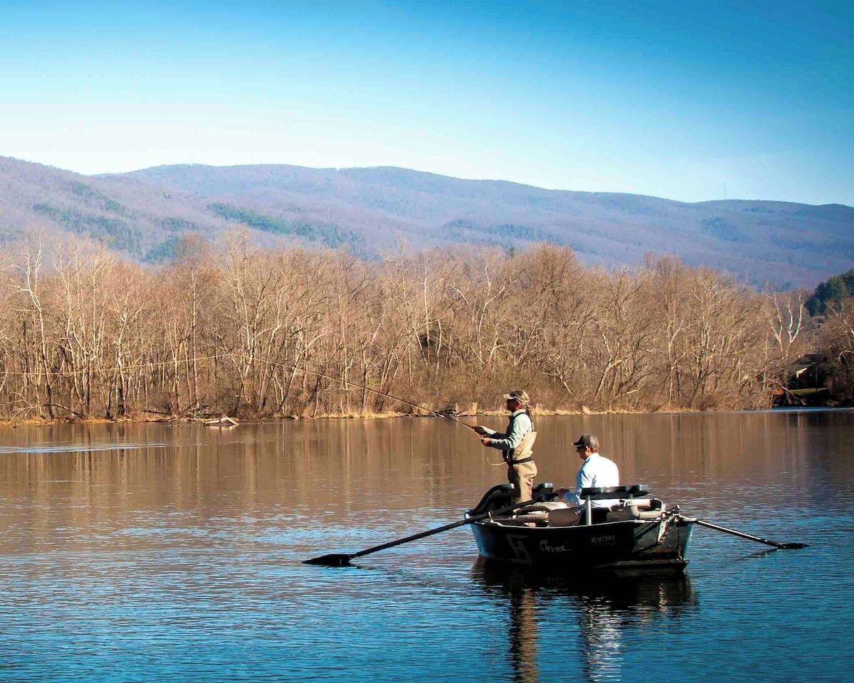 A calm lake with a boat carrying two people fishing, surrounded by leafless trees and distant hills.