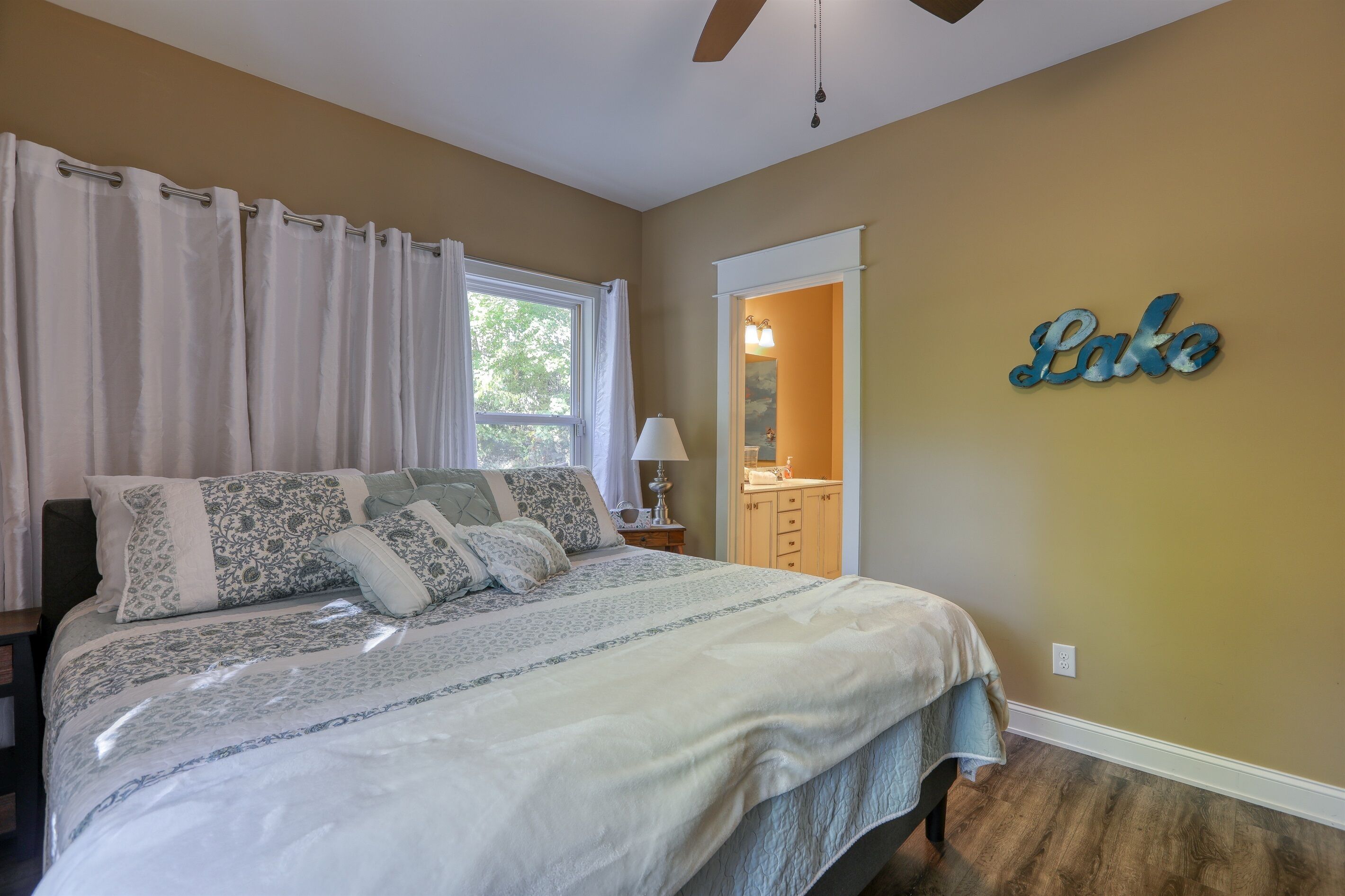 Un dormitorio con cama, mesita de noche, lámpara, ventana con cortinas, espejo y un cartel en la pared que dice "Lake".