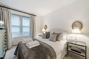 Habitación de hotel con cama, dos lámparas de noche, ventana con cortinas, espejo en la pared y mesita de noche con lámpara.