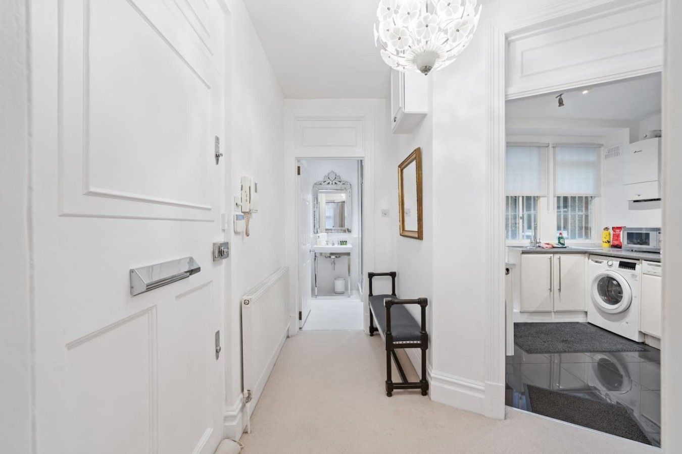 A hallway leading to a bathroom with a mirror, a bench, and a chandelier.