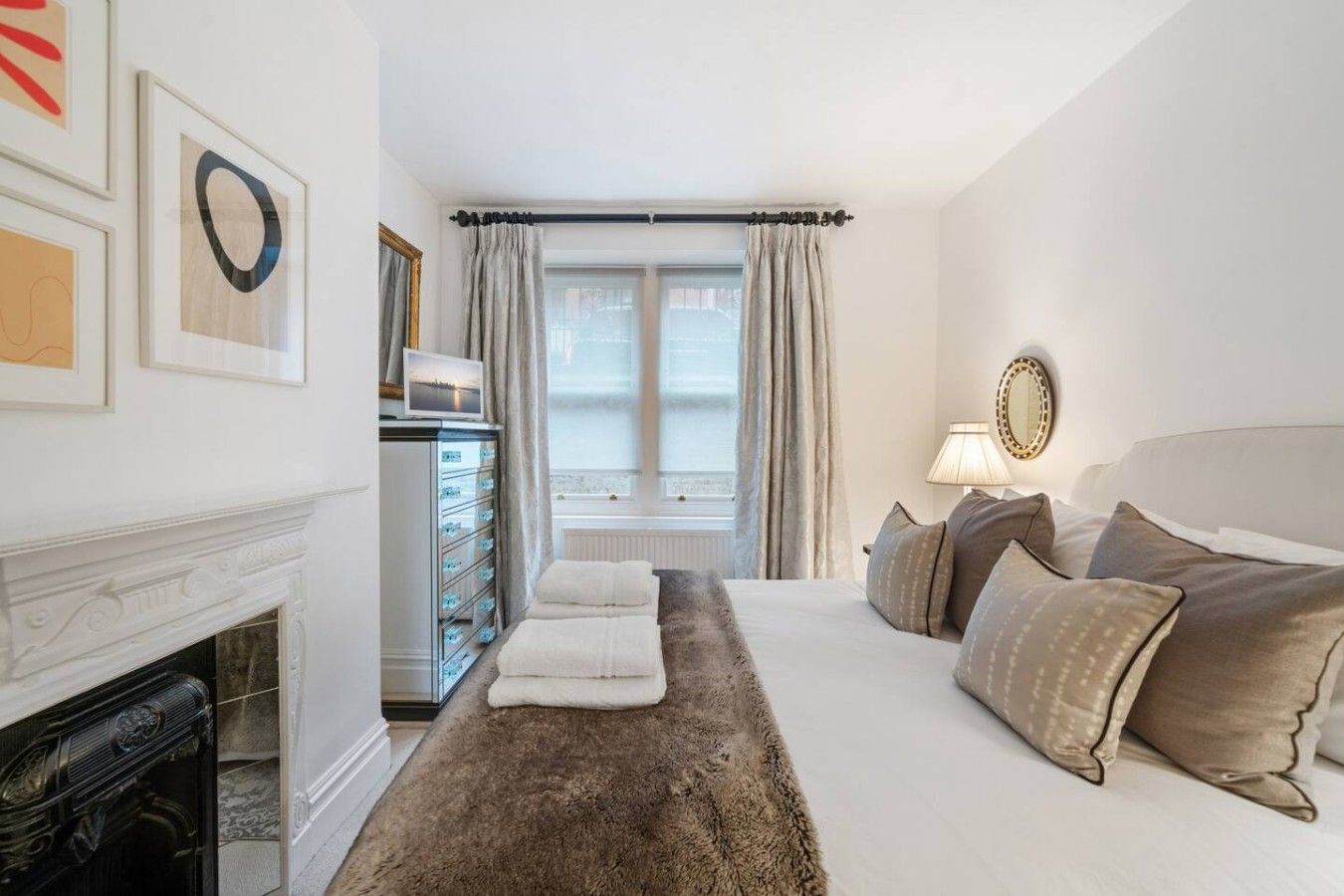 A hotel room with a fireplace, a bed with multiple pillows, a dresser with drawers, a window with curtains, and wall art.