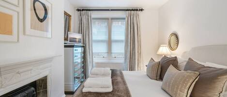 A hotel room with a fireplace, a bed with multiple pillows, a dresser with drawers, a window with curtains, and wall art.