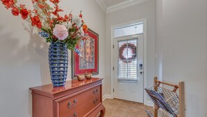A hallway with a wooden dresser, a vase of flowers, a framed picture, and a door with a wreath. - Spacious Zachary Home: 12 Mi to Baton Rouge! (Zachary)