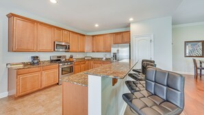 A modern kitchen with wooden cabinets, granite countertops, stainless steel appliances, and a bar area with leather barstools. - Spacious Zachary Home: 12 Mi to Baton Rouge! (Zachary)