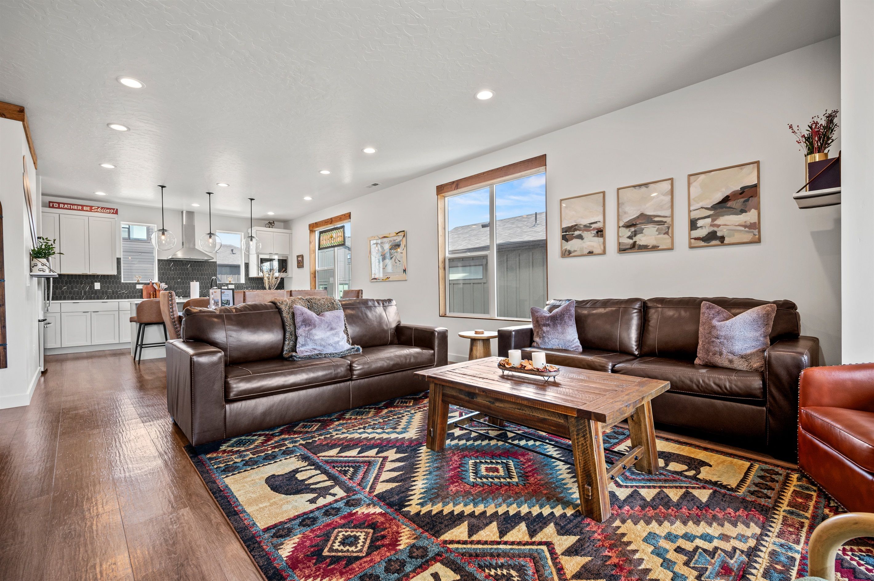 A spacious living room with a brown leather sofa, a coffee table, and a patterned rug. There is a kitchen in the background with white cabinets and a large window.