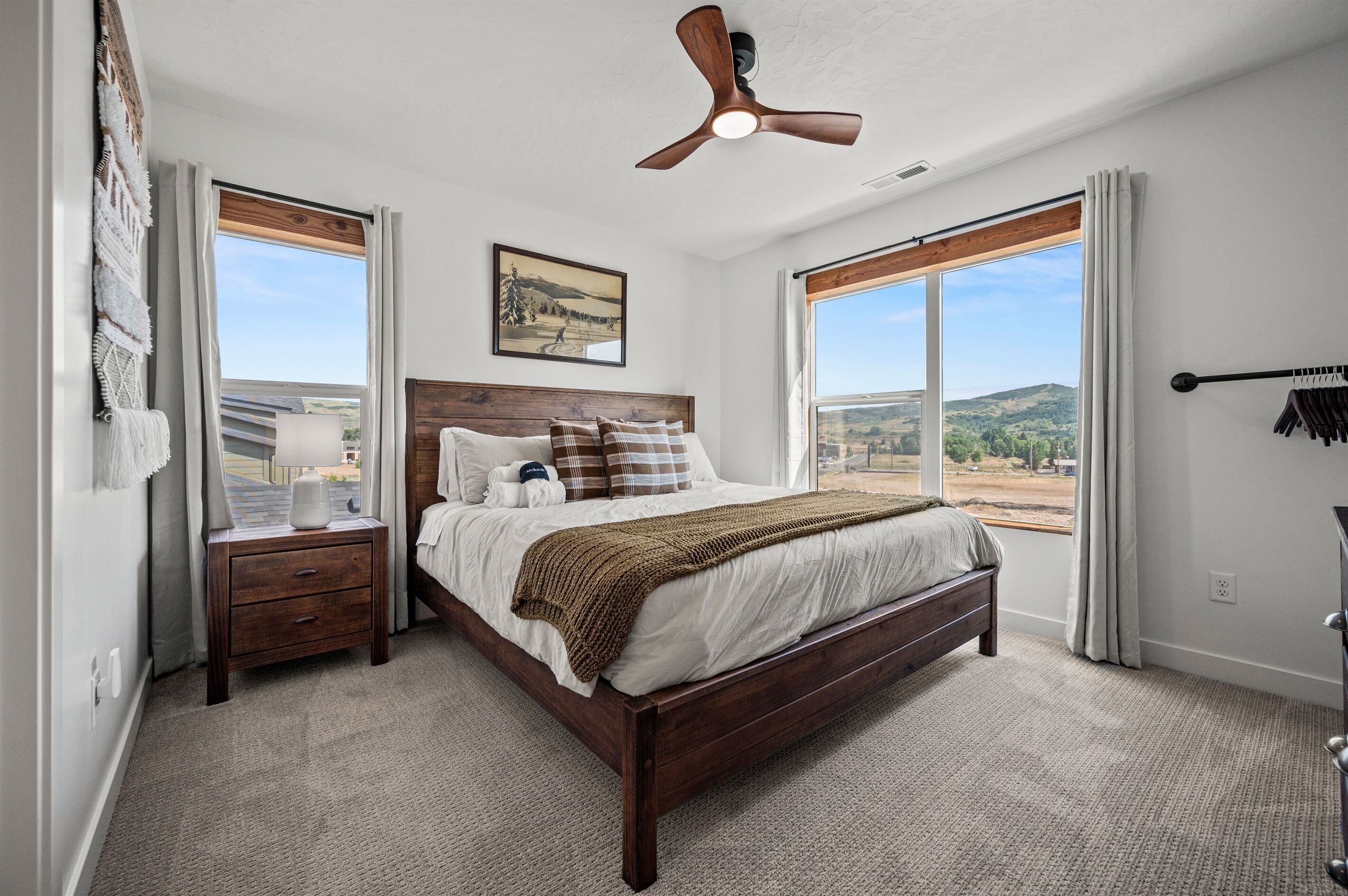 A bedroom with a large bed, a nightstand, a ceiling fan, and a window with a view of the outdoors.