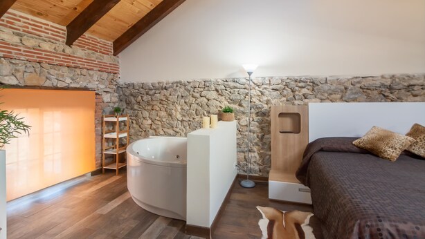 Een slaapkamer met een stenen muur, houten balken, een groot bed, een nachtkastje en een ingebouwde bank.