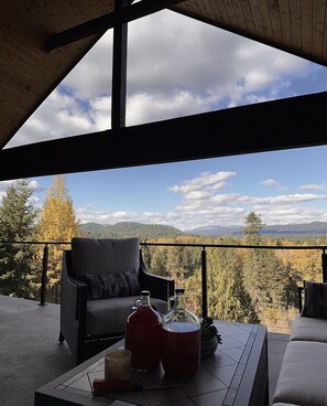 A balcony with a view of a forest and mountains, a wicker chair, a table with a bottle and a mug, and a wooden ceiling.