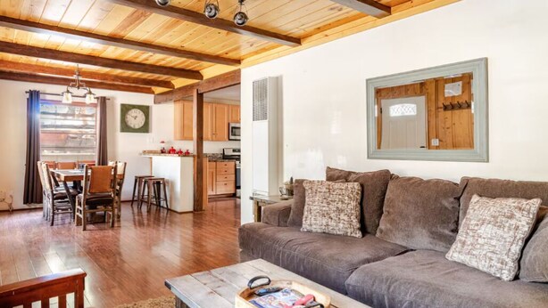 A cozy living room with a wooden ceiling, a sofa with cushions, a coffee table with books, and a dining area with chairs and a table.