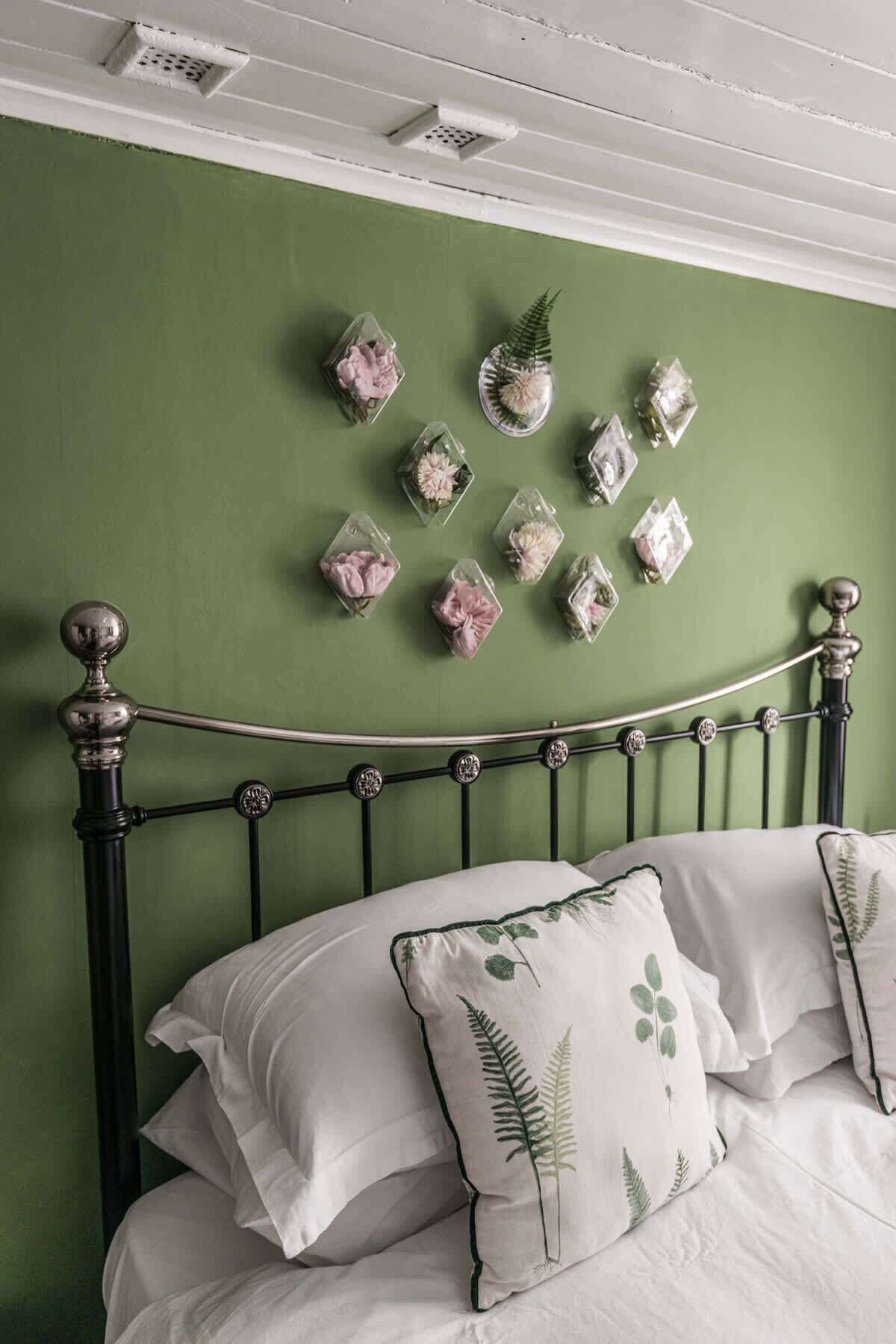 A bed with white bedding and green leaf-patterned pillows, a metal headboard, and decorative glass containers with flowers on a green wall.