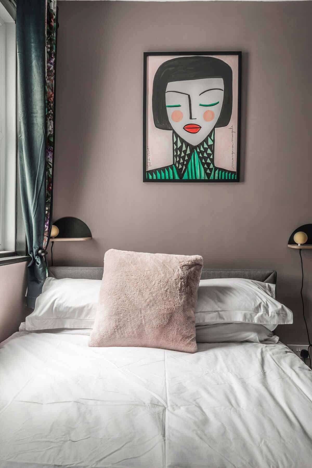 Ein Bett mit weißer Bettdecke und einem rosa Kopfkissen, ein gerahmtes Kunstwerk an der Wand und ein Fenster mit Vorhängen.