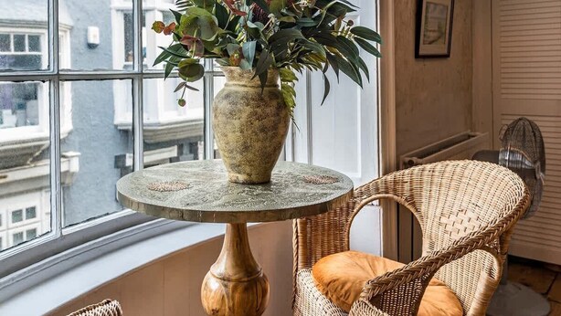 A room with a large window, wicker chairs, a round table with a vase of plants, and a wall-mounted fan. - Old Post Office - Large house with games room +bar (Ramsgate)