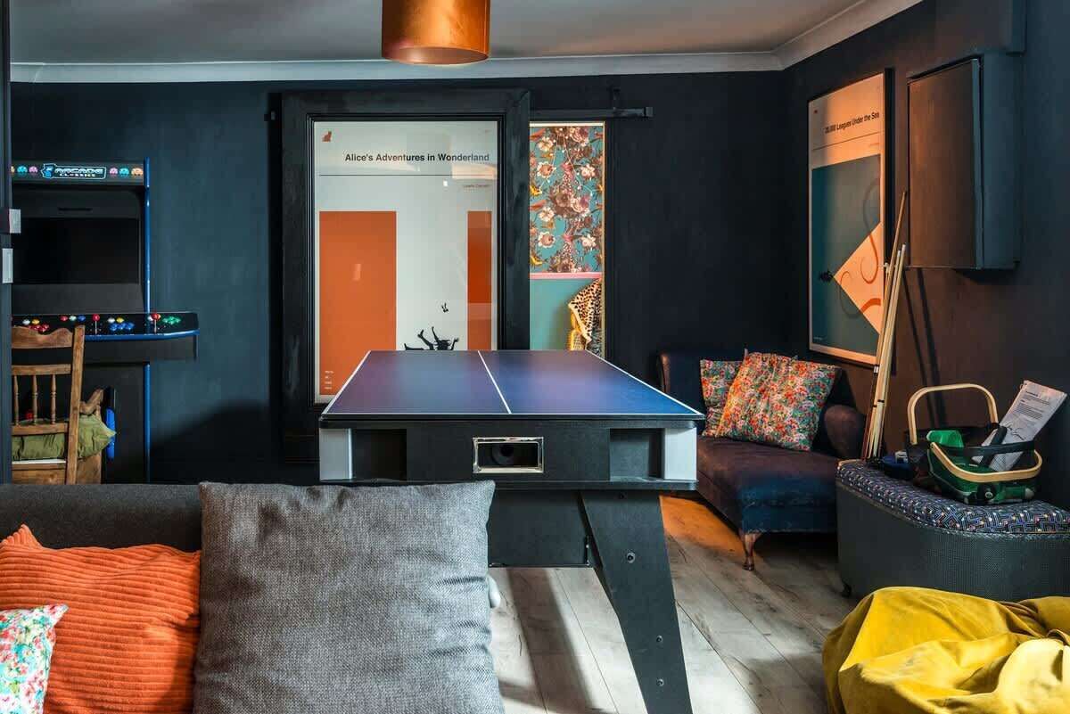 A room with a ping pong table, a sofa, a small table with a laptop, and a wall with a poster.