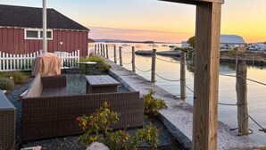 A waterfront patio with wicker furniture, a covered table, and a view of boats on the water.