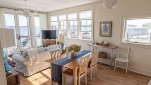 A spacious living room with a wooden dining table, white sofas, and a TV. Large windows offer a view of the sea.