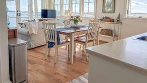 A bright, sunlit room with a wooden dining table, white chairs, and a TV on a stand.