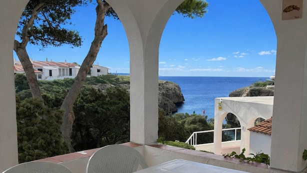 Une table blanche avec des chaises, placée dans un espace extérieur abrité, offrant une vue sur la côte et un ciel dégagé.