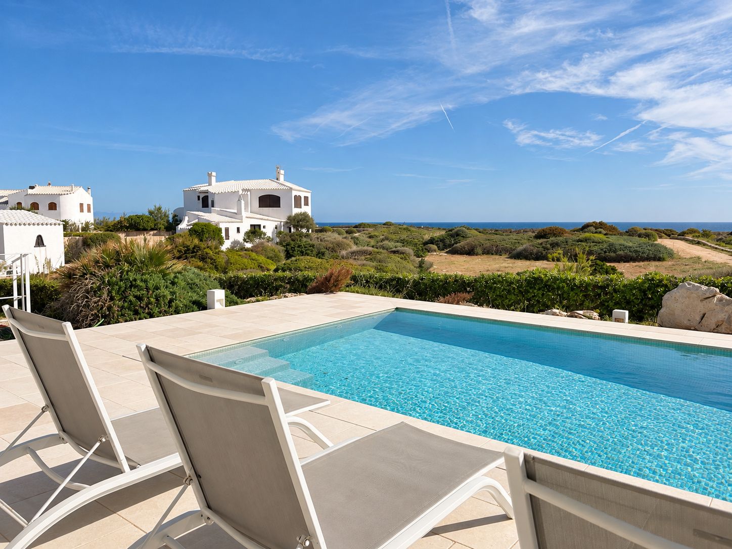 Una piscina con sillas de descanso, un edificio blanco y vistas al mar.