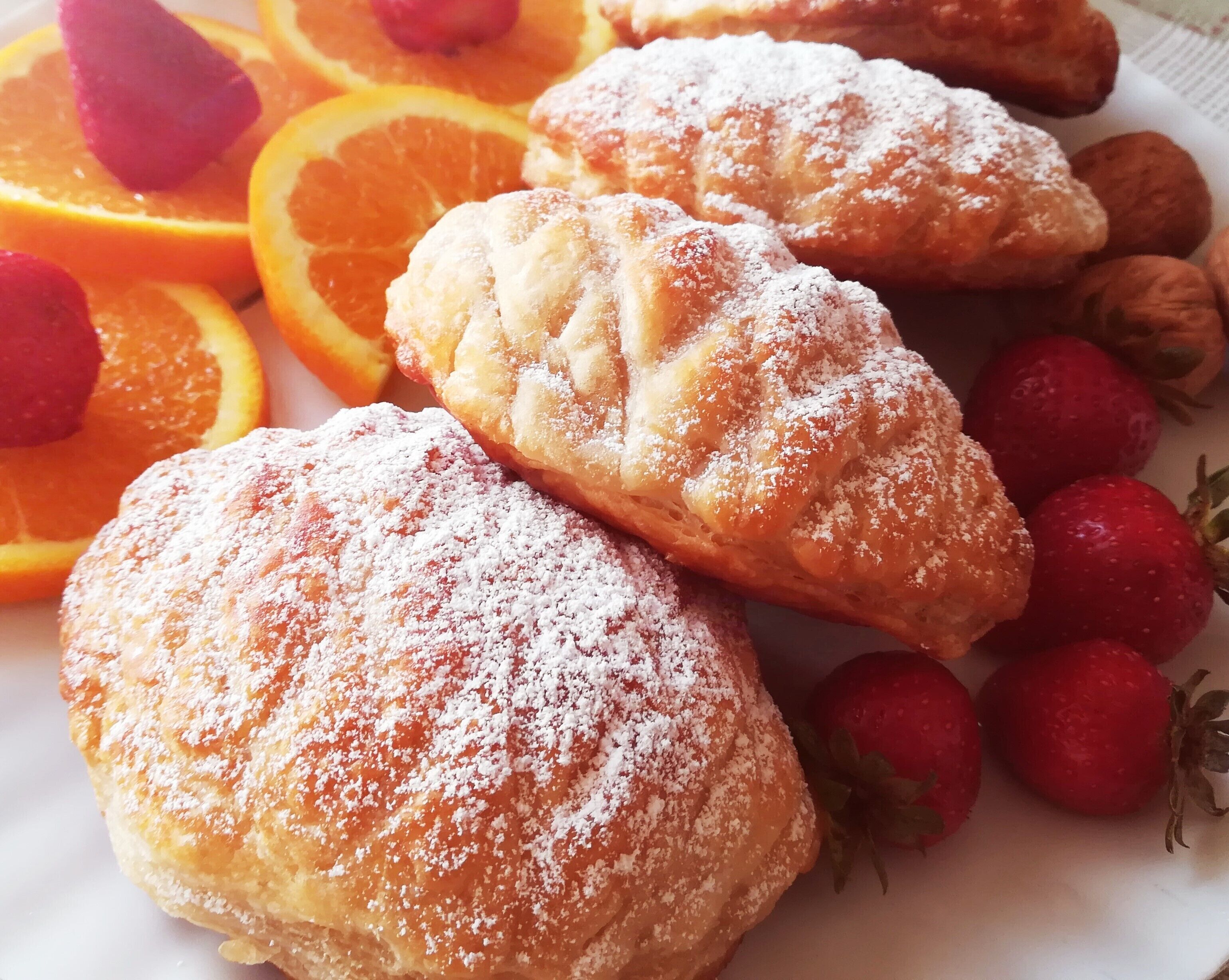 Pastries dusted with powdered sugar, accompanied by strawberries and orange slices.
