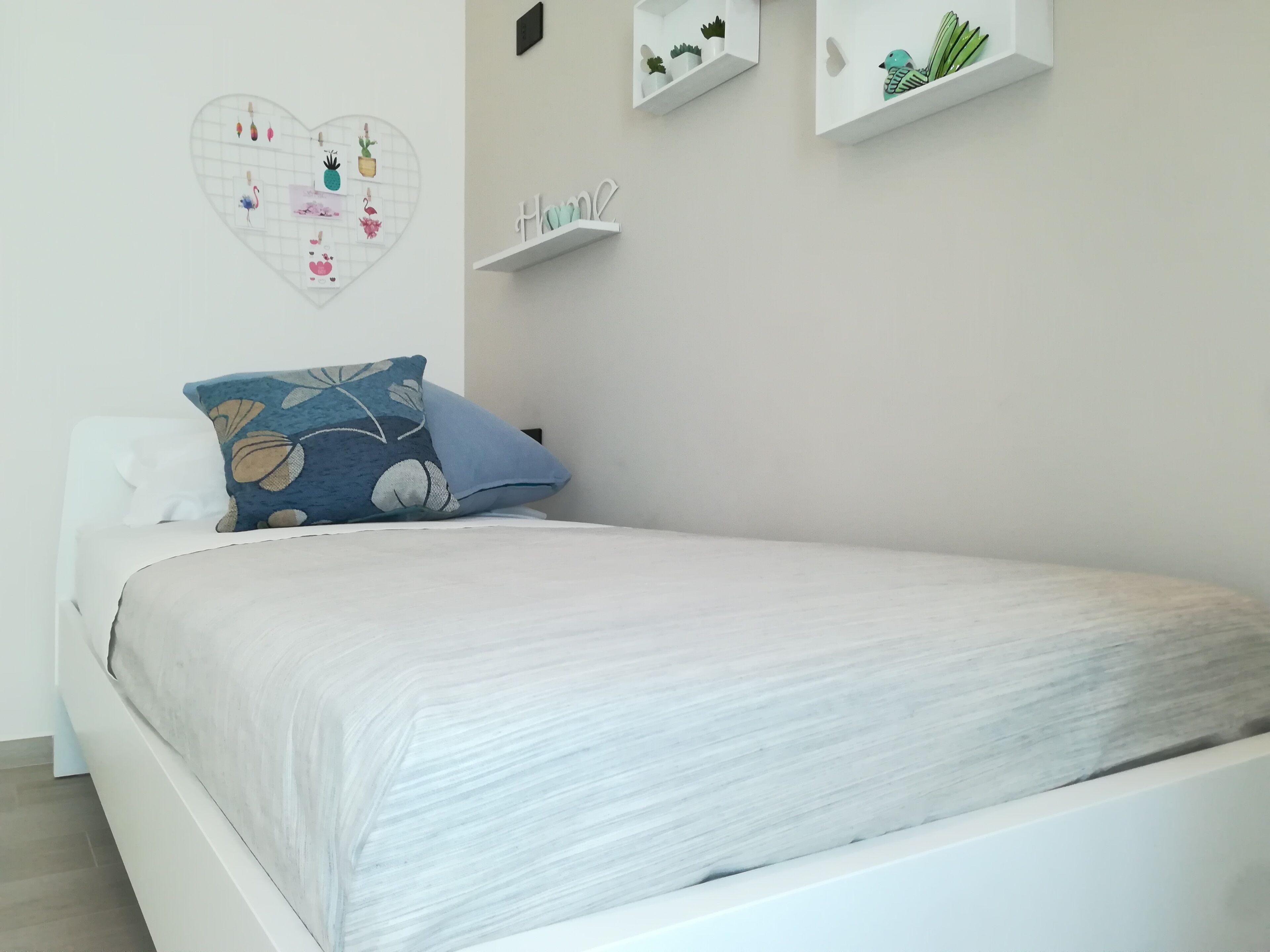 A neatly made bed with a white mattress and blue floral pillows, a heart-shaped photo frame with various decorations, and a small shelf with a 'Home' sign and potted plants.