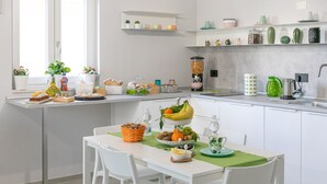 Une cuisine moderne avec des meubles blancs, un coin repas avec des chaises blanches, et divers aliments et appareils de cuisine disposés sur les plans de travail.