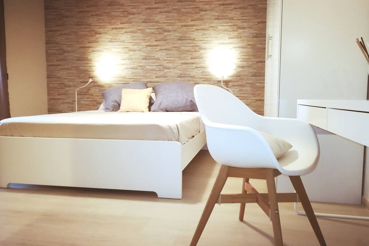 Un dormitorio moderno con cabecera de madera, cama blanca y un escritorio blanco con silla.