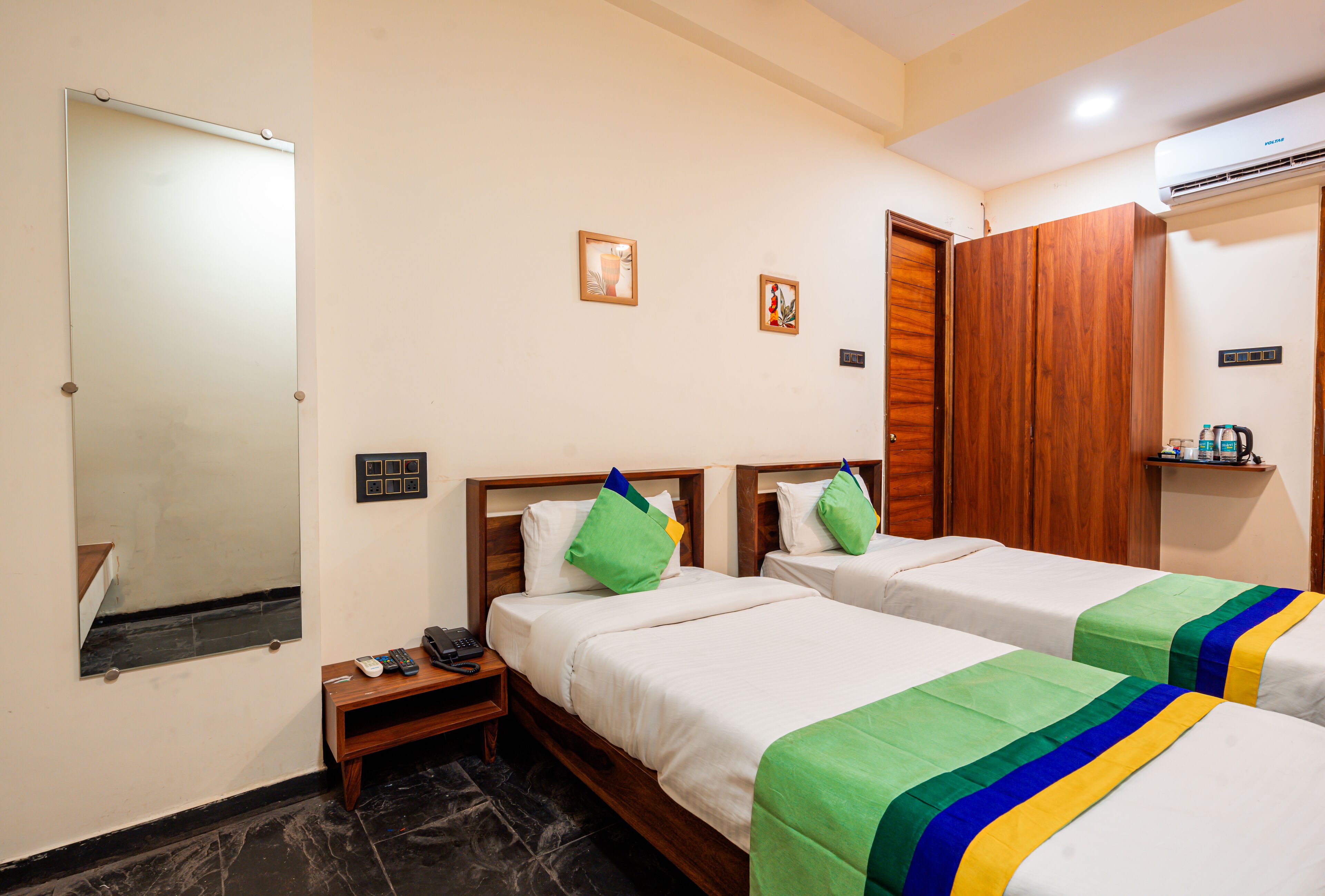 A hotel room with two beds, a wooden wardrobe, a mirror, a small table with a phone and remote control, and a wall-mounted air conditioner.