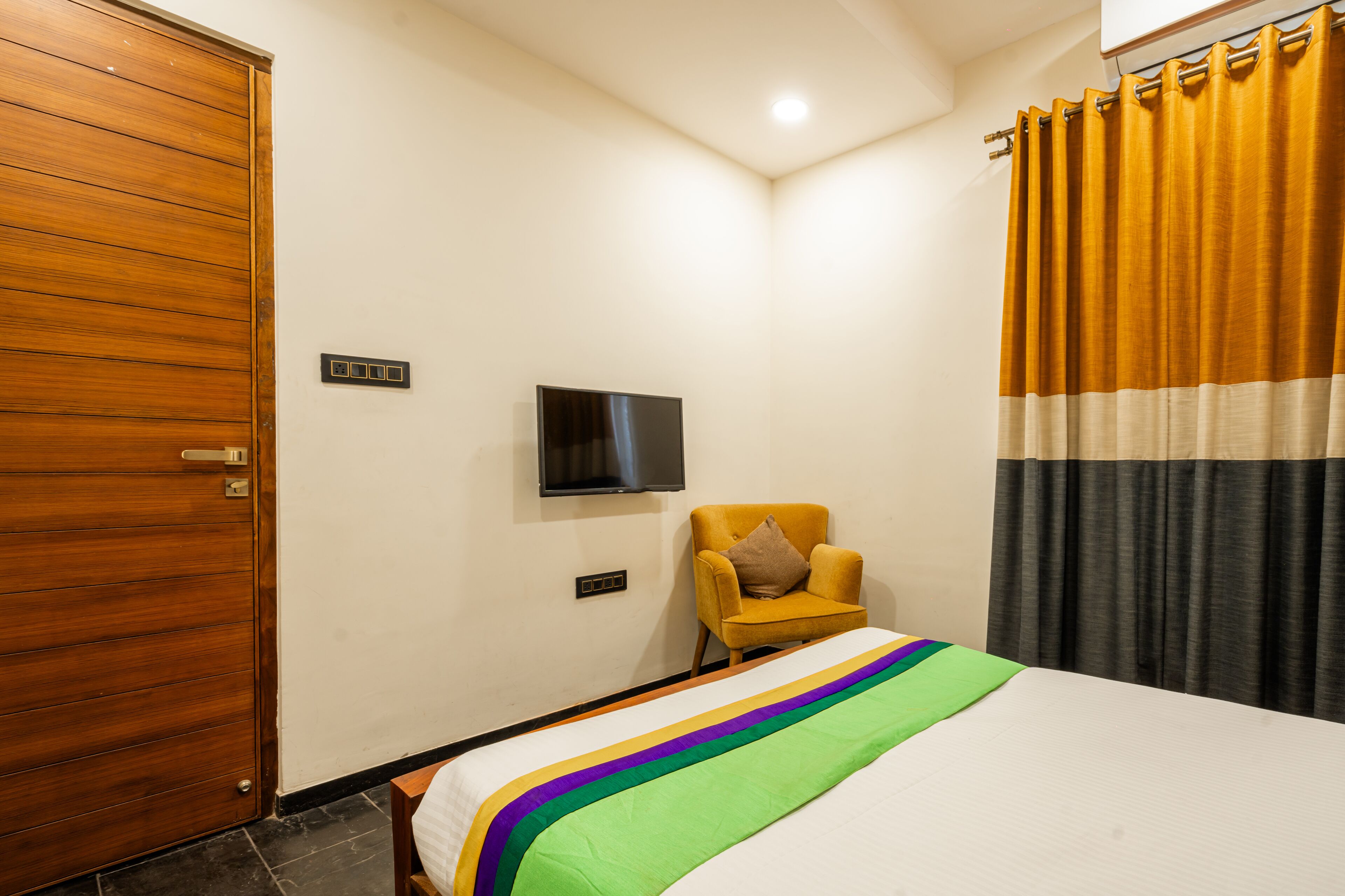 Habitación de hotel con puerta de madera, cama con colcha colorida, sillón pequeño, televisor de pantalla plana montado en la pared y cortina con estampado a rayas.