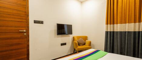 Habitación de hotel con puerta de madera, cama con colcha colorida, sillón pequeño, televisor de pantalla plana montado en la pared y cortina con estampado a rayas.