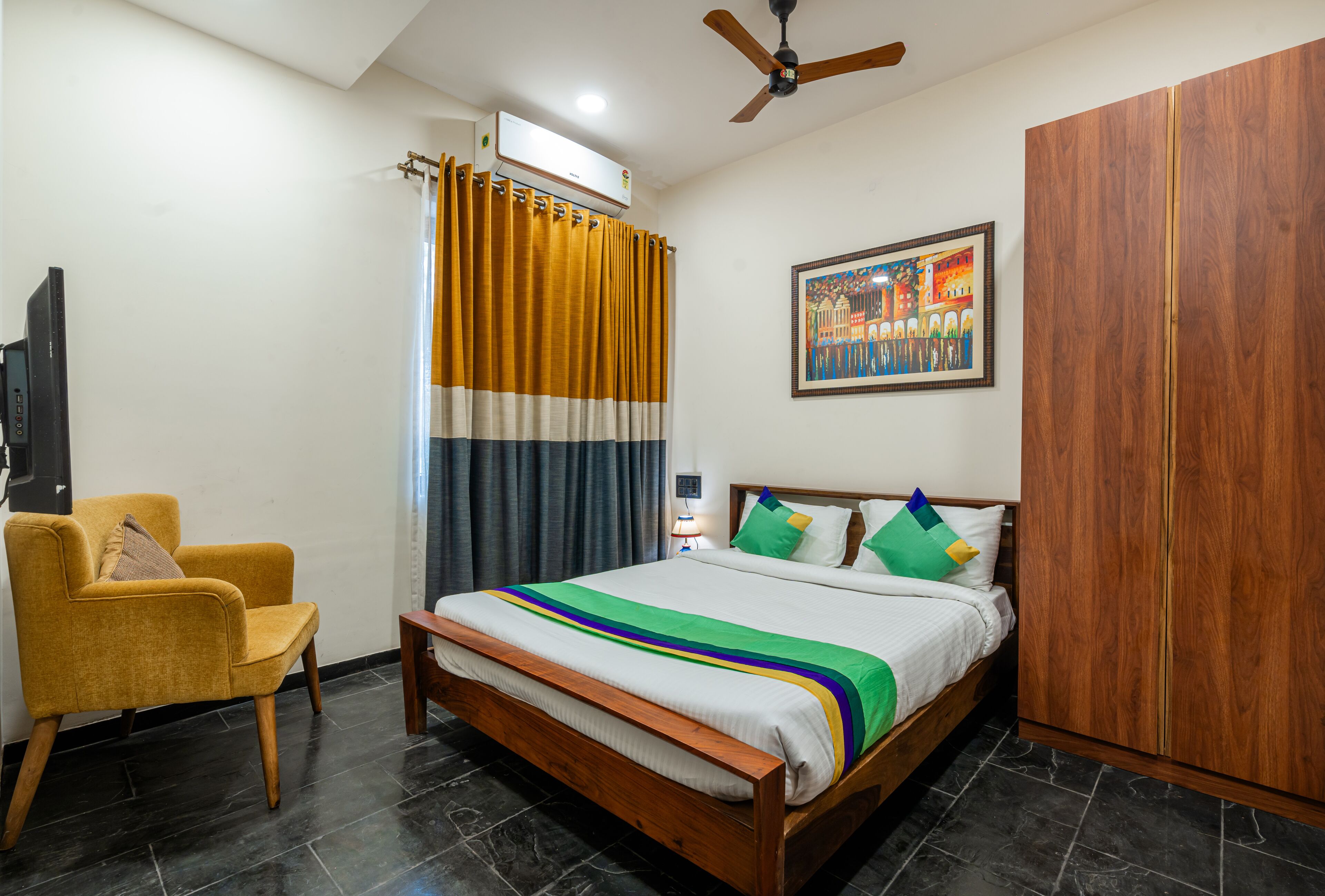 Un dormitorio con una cama, un armario de madera, un sillón amarillo, un ventilador de techo y una ventana con cortinas.