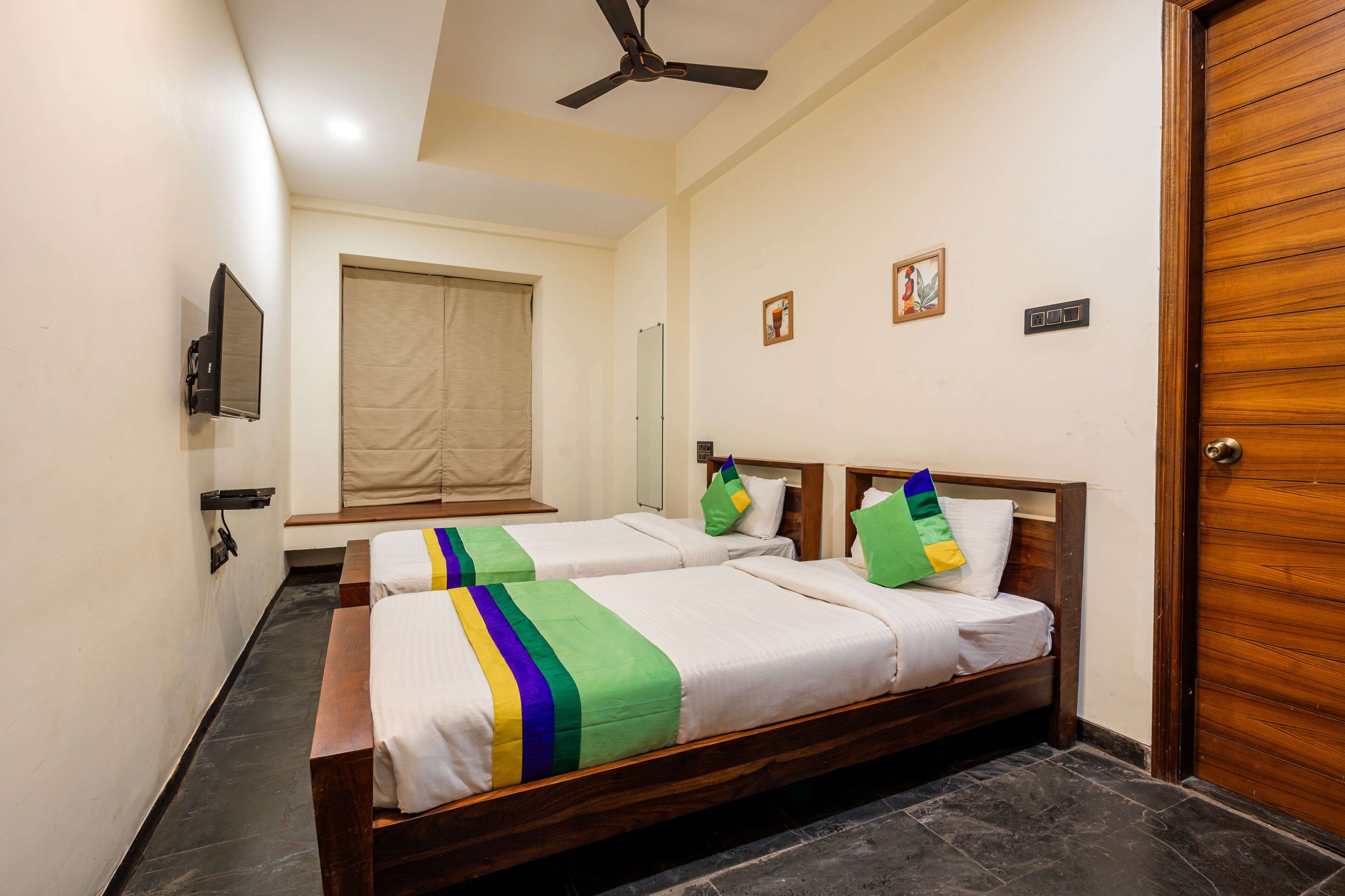 A hotel room with two single beds, a ceiling fan, a flat-screen TV, a window with a beige blind, and a wooden door.