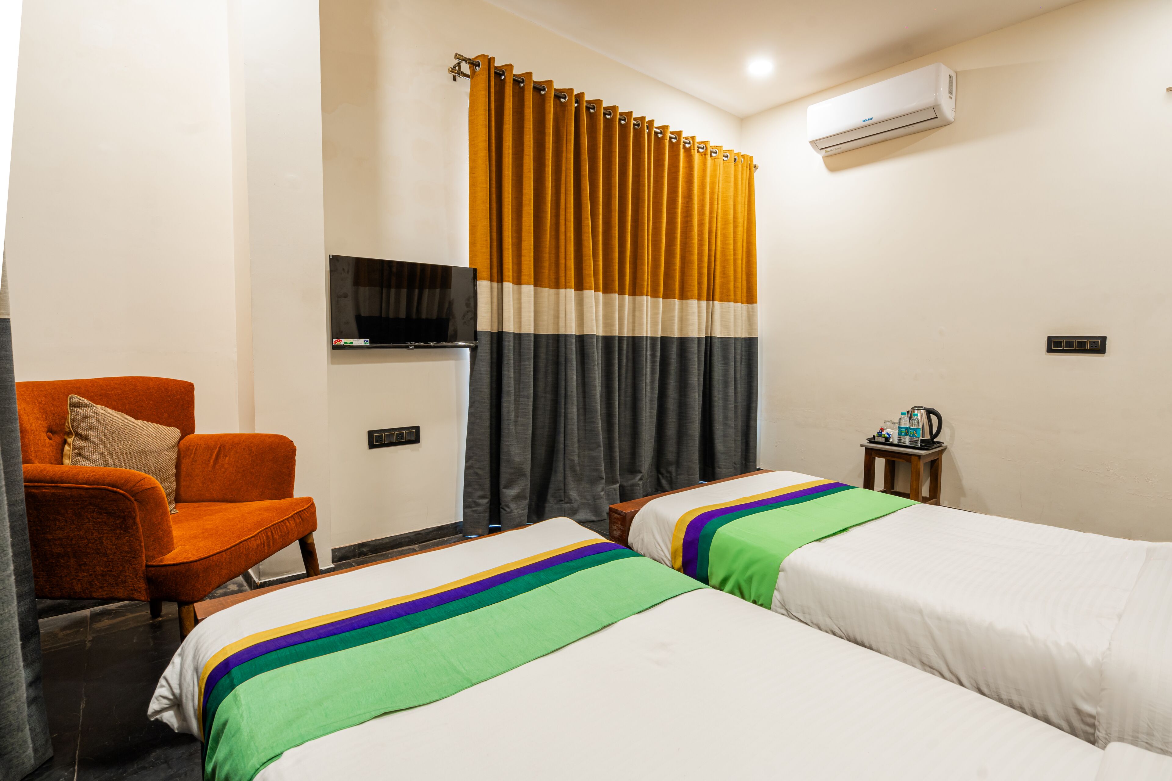 Una habitación de hotel con dos camas, un sillón naranja, un televisor y una mesita con un hervidor.