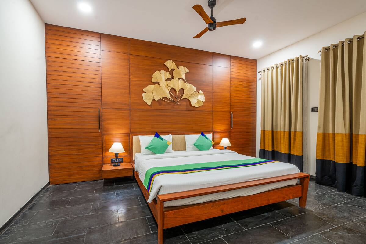 A bedroom with a wooden bed frame, a ceiling fan, a nightstand with a lamp, and a wall-mounted artwork.