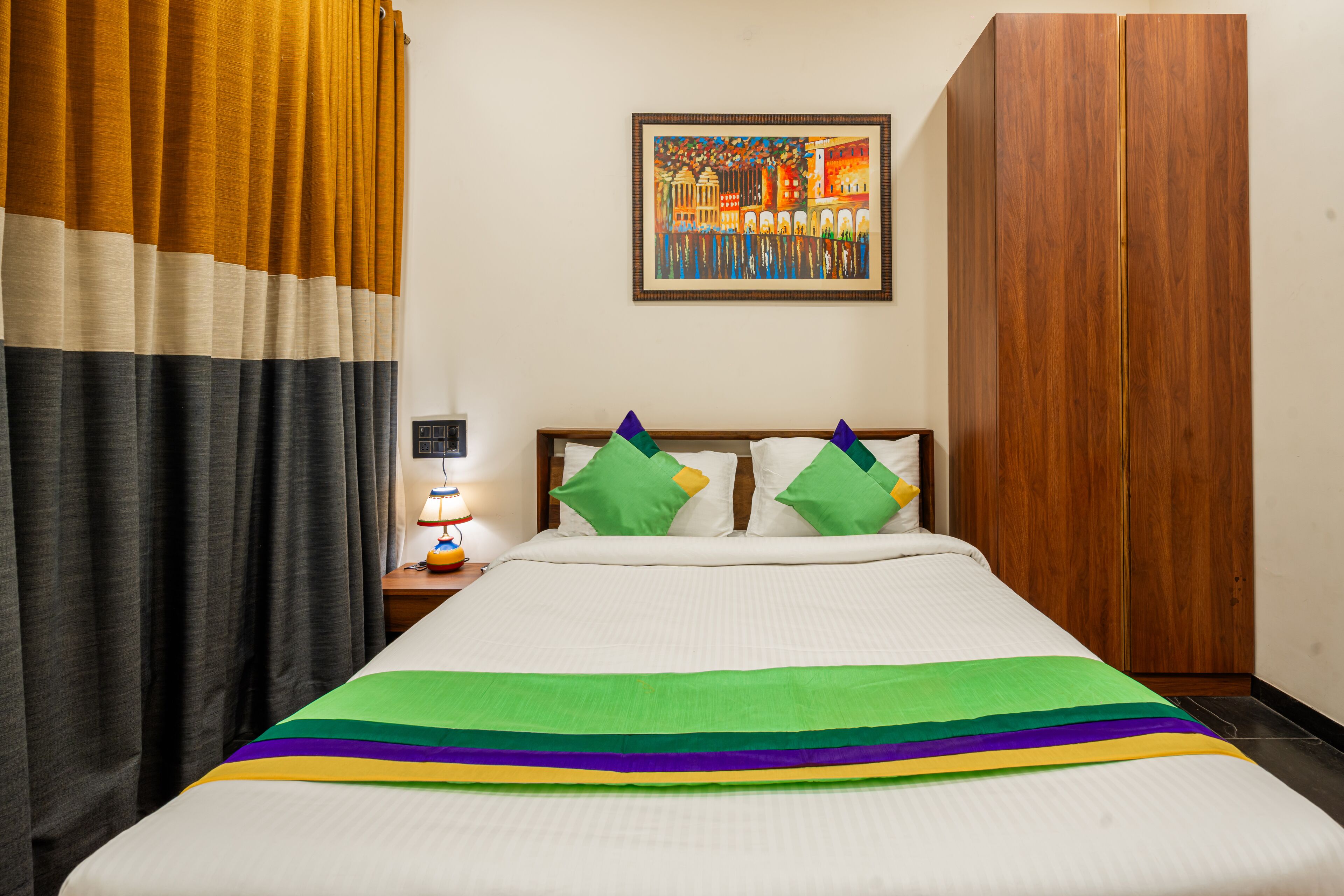 Een hotelkamer met een bed, gordijnen, een nachtkastje met een lamp, een schilderij aan de muur en een kledingkast.