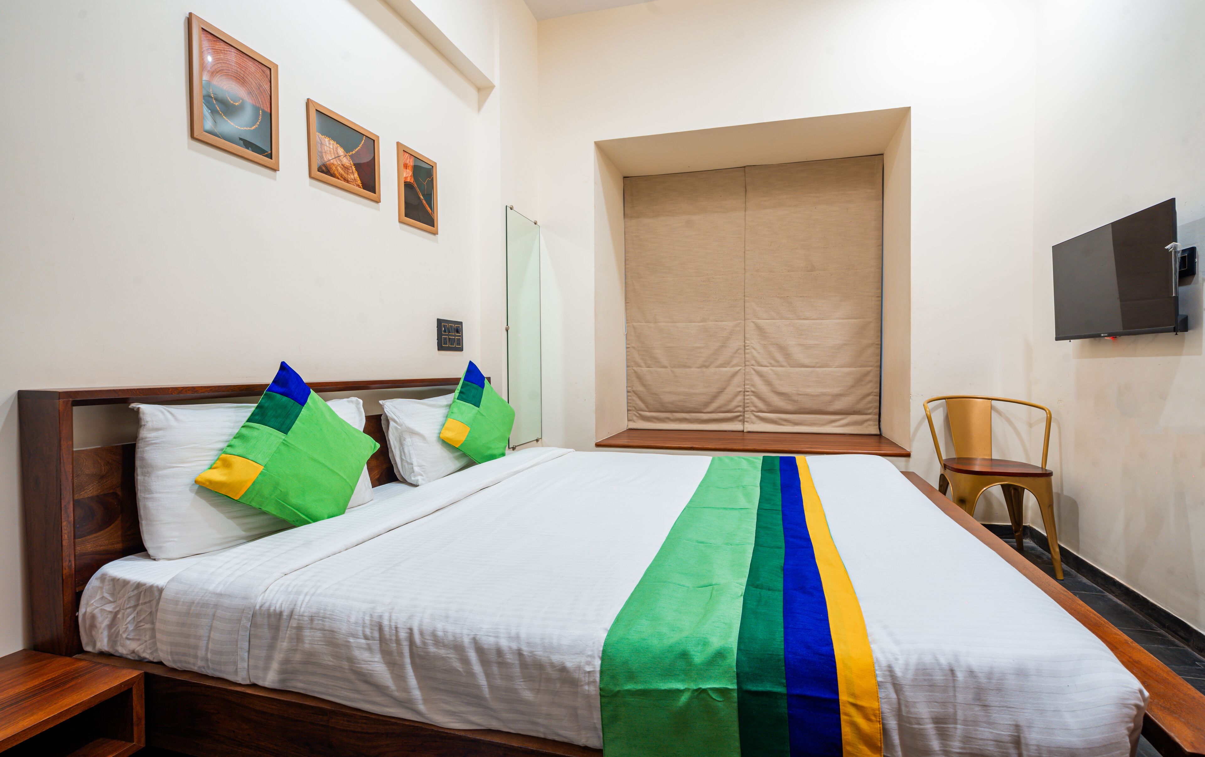 Une chambre d’hôtel avec un grand lit, une tête de lit en bois, une chaise, un téléviseur et trois tableaux encadrés accrochés au mur.