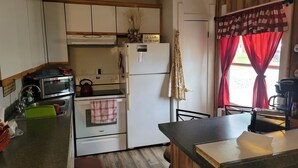 Cocina compacta con microondas, estufa y refrigerador. Hay un lavamanos con un paño verde y una taza roja. La cocina tiene armarios de madera y una ventana con cortinas rojas.