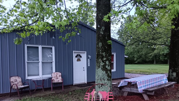 Un pequeño edificio de una sola planta con puerta y ventanas blancas, un porche con dos sillas y una mesa de picnic cubierta con mantel a cuadros.