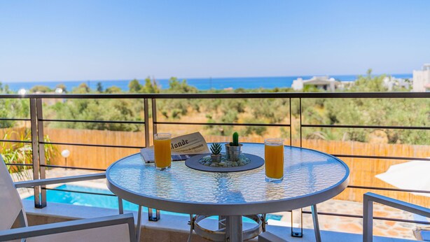 Et rundt bord med to glas appelsinjuice, en lille potteplante og en menu på en balkon med udsigt over en pool og havet.