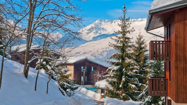 Una cabaña de madera con balcón, rodeada de árboles nevados y montañas al fondo.