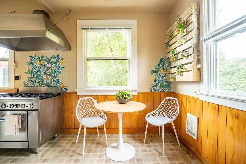 A kitchen with a gas stove, oven, and a window with a view of greenery. There are two white chairs and a small table with a plant.