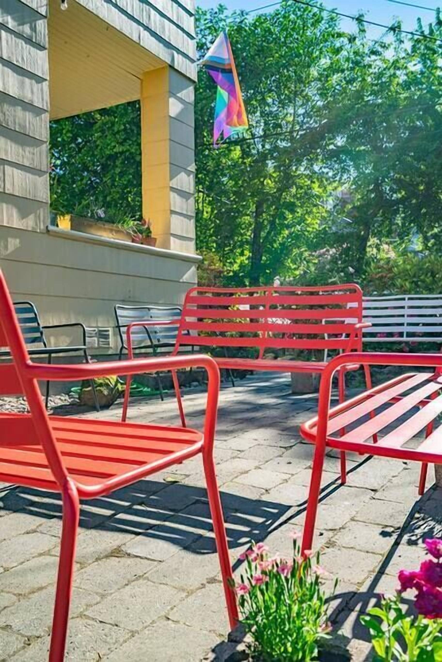 An outdoor seating area with red metal chairs and a white bench, surrounded by greenery and a building with a yellow window frame.