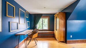 A room with blue walls, wooden flooring, a wooden desk, a chair, and a window with a view of greenery. - Charming Room in Historic BnB - Rm6 (Seattle)