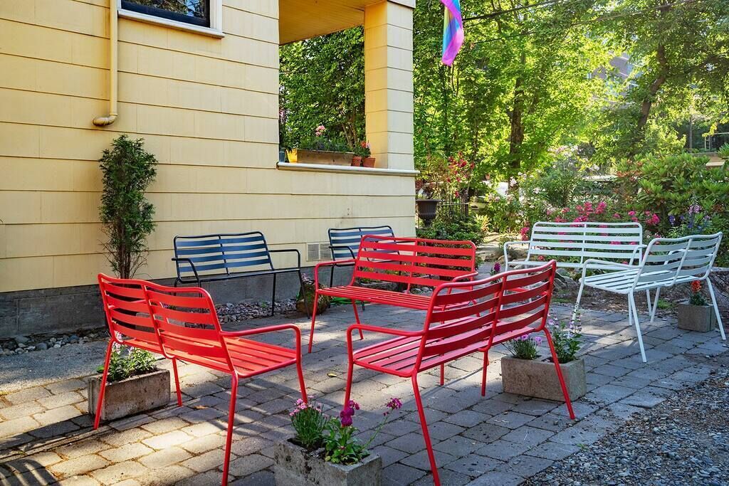 An outdoor seating area with red and white metal chairs and tables, surrounded by potted plants and a yellow building.