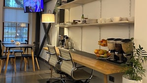 Un espacio de oficina moderno con una larga mesa de madera, sillas de oficina, un televisor montado en la pared y estanterías con libros y utensilios de cocina.