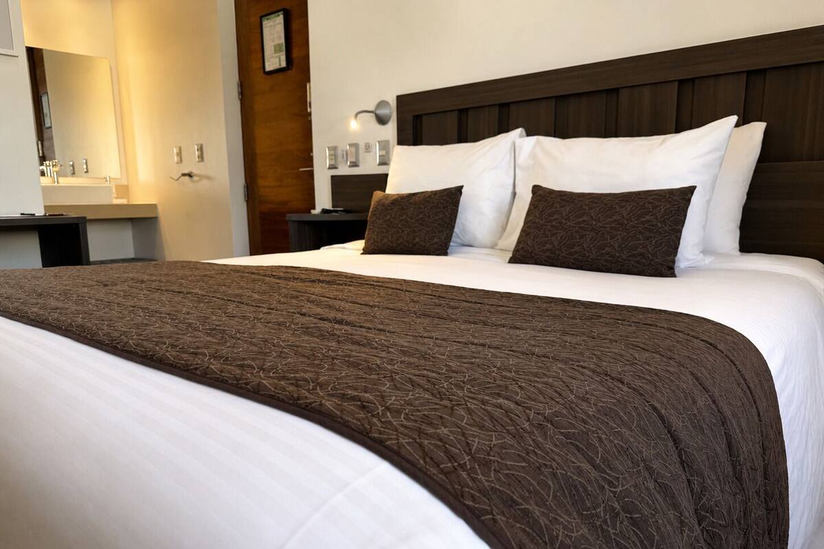 A hotel room with a large bed, a wooden headboard, a nightstand, a mirror, and a bathroom visible in the background.